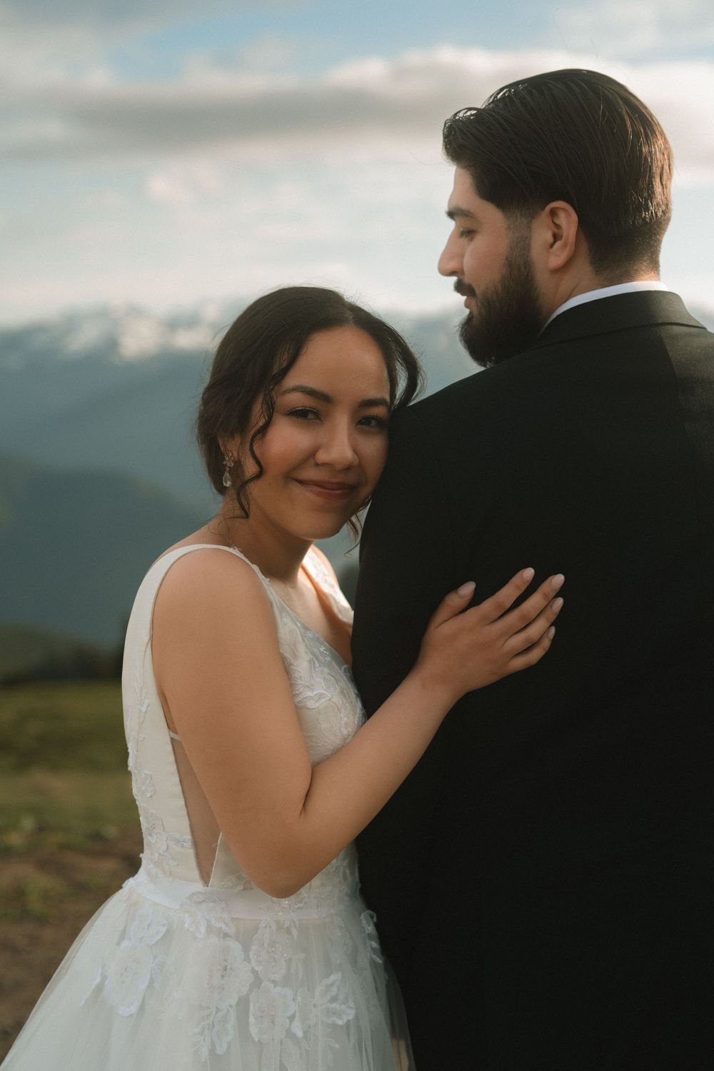 Bride in white lace gown embracing groom in black suit with mountain backdrop.