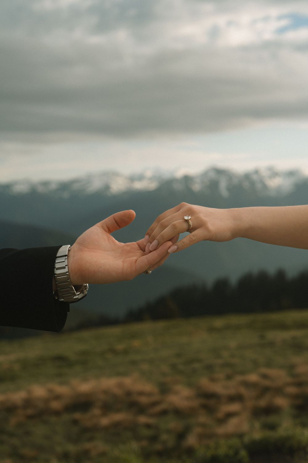 Bride and groom reach for each other's hands with mountains and cloudy sky in the background.