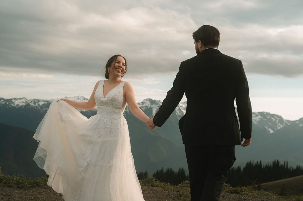 A bride in a white gown and groom in a black suit hold hands on a mountain with snowy peaks and cloudy skies.