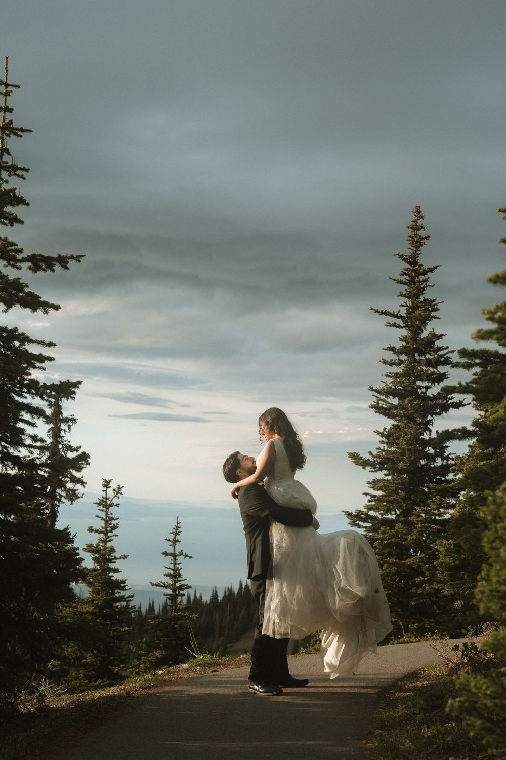 Groom lifts bride in white wedding dress on a mountain road surrounded by evergreen trees at dusk.