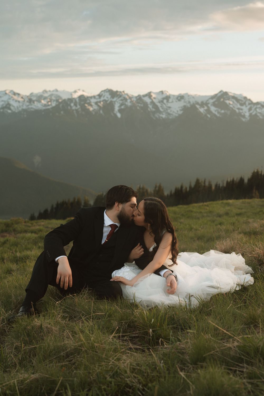 Couple shares a romantic kiss while seated on a grassy mountain meadow with snow-capped peaks behind them.
