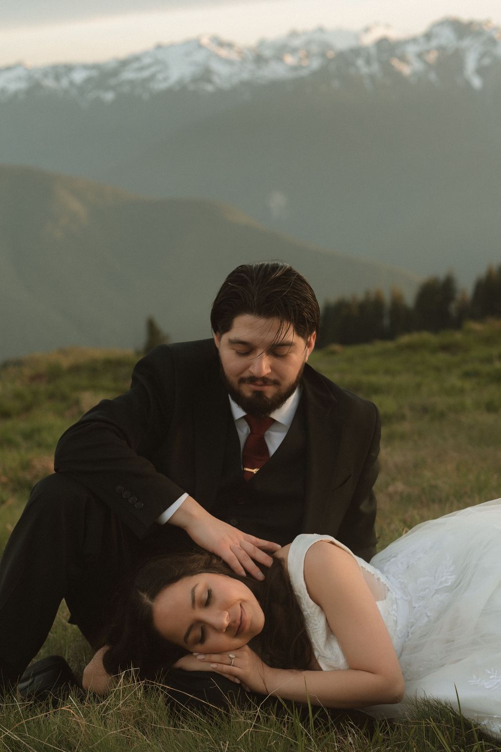 Bride and groom in wedding attire resting on mountain meadow with snow-capped peaks backdrop at dusk.