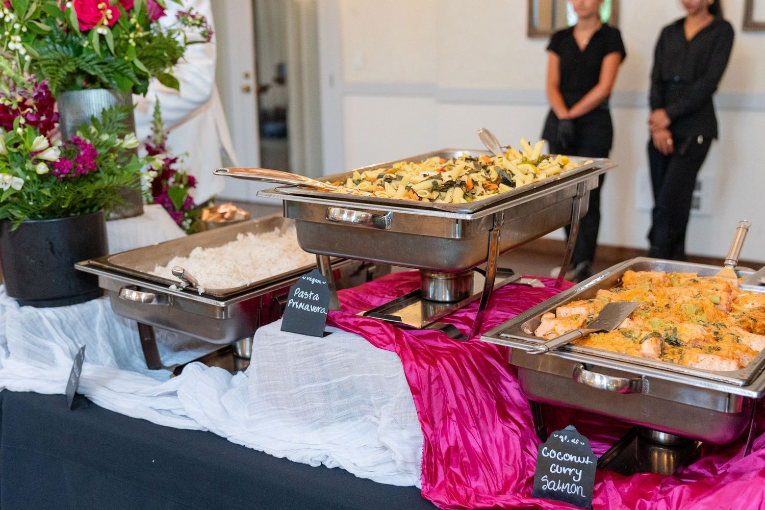 Elegant catered buffet table with chafing dishes filled with colorful food, pink satin tablecloth, and floral arrangement.