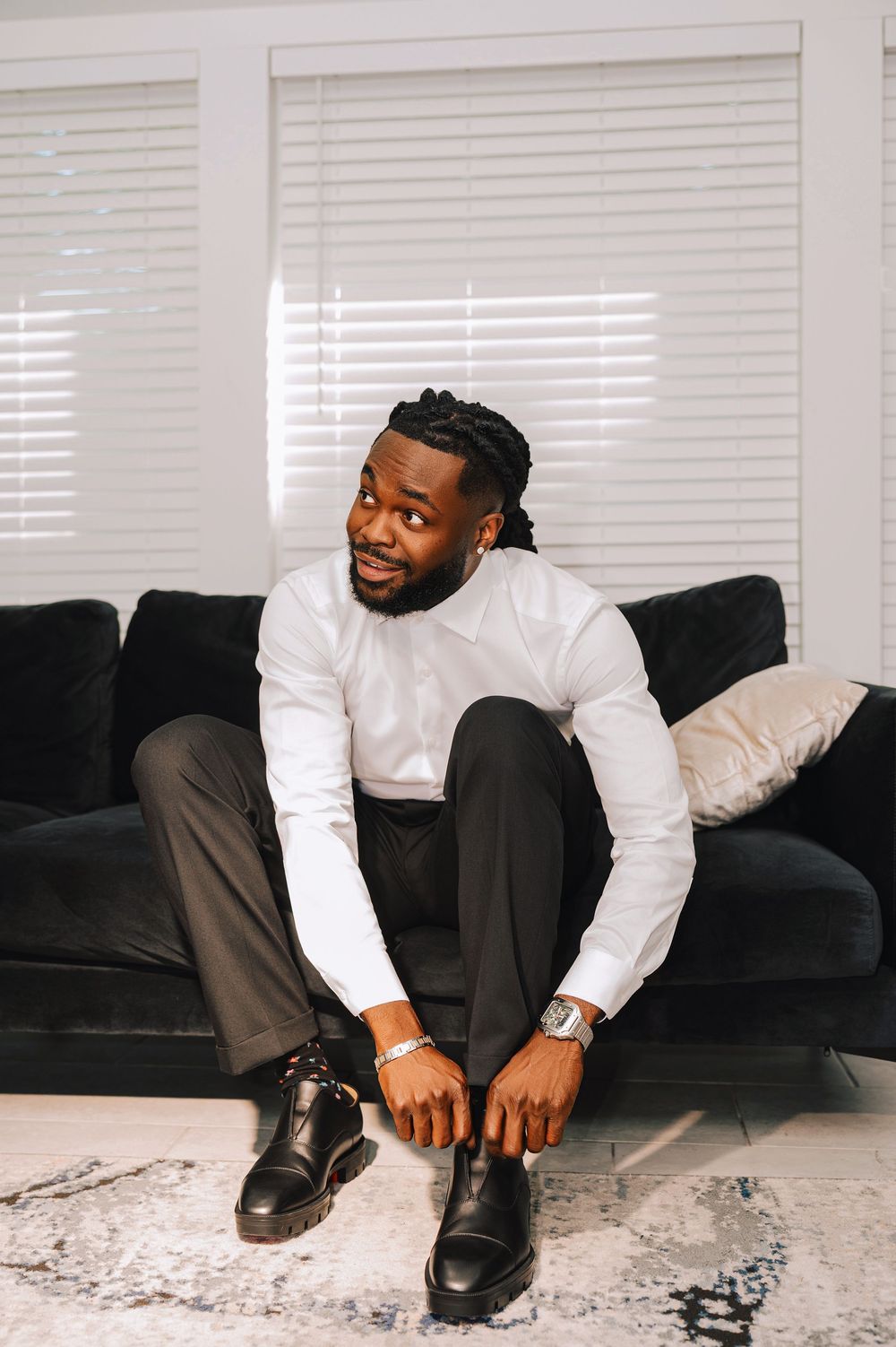 A well-dressed Black man in a white shirt and dark trousers sits on a black sofa, putting on black dress shoes.
