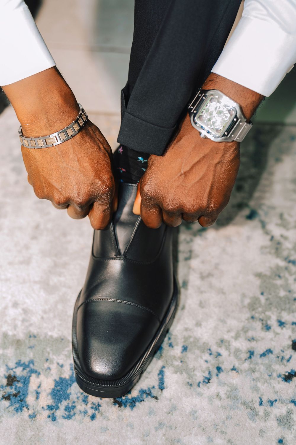 Man in dress pants putting on black leather dress shoes while wearing a silver watch and bracelet.