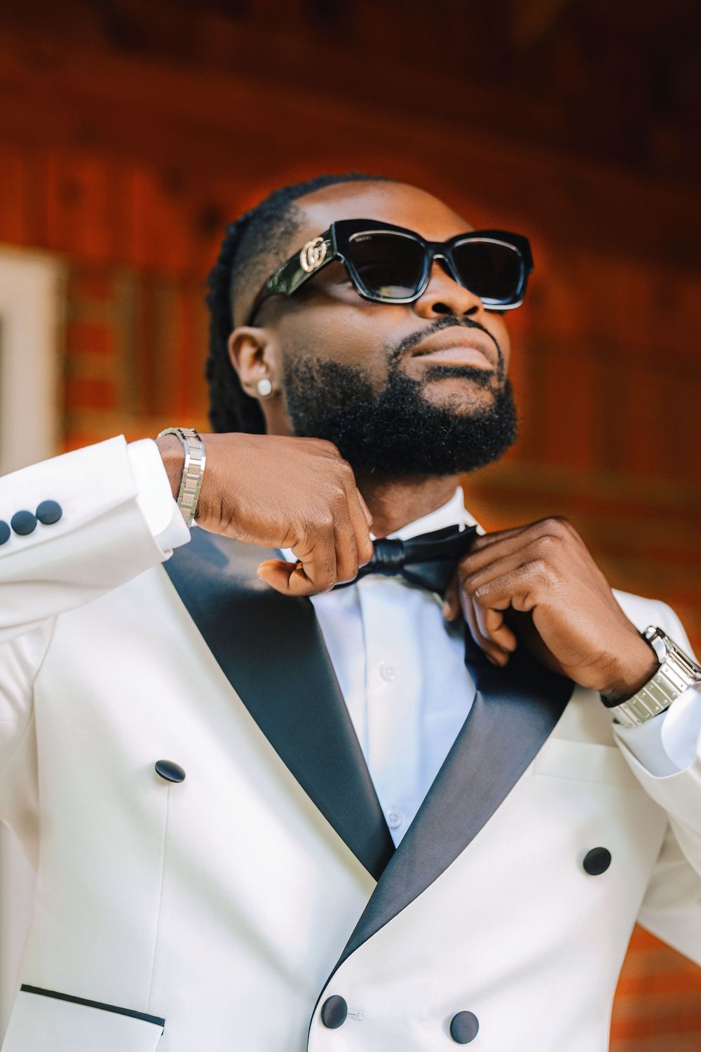 Stylish man in white tuxedo with black lapels adjusting bow tie, wearing sunglasses and watch, elegant formal attire.