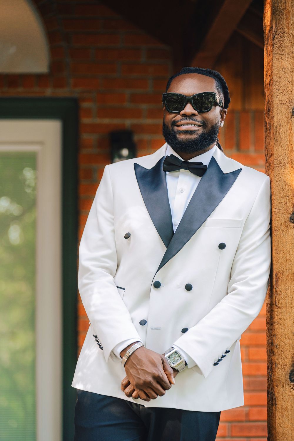 A well-dressed Black man in a white double-breasted tuxedo jacket, bow tie, and sunglasses smiles while leaning against a brick wall.
