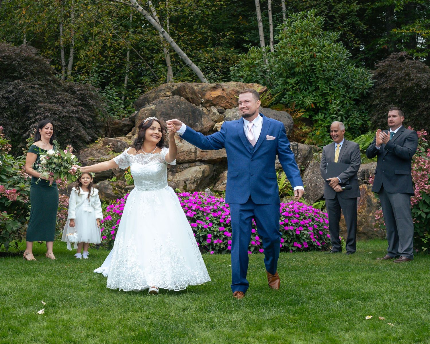 Bride and groom celebrate outdoors at garden wedding with bridal party, surrounded by lush greenery and pink flowers at outdoor wedding