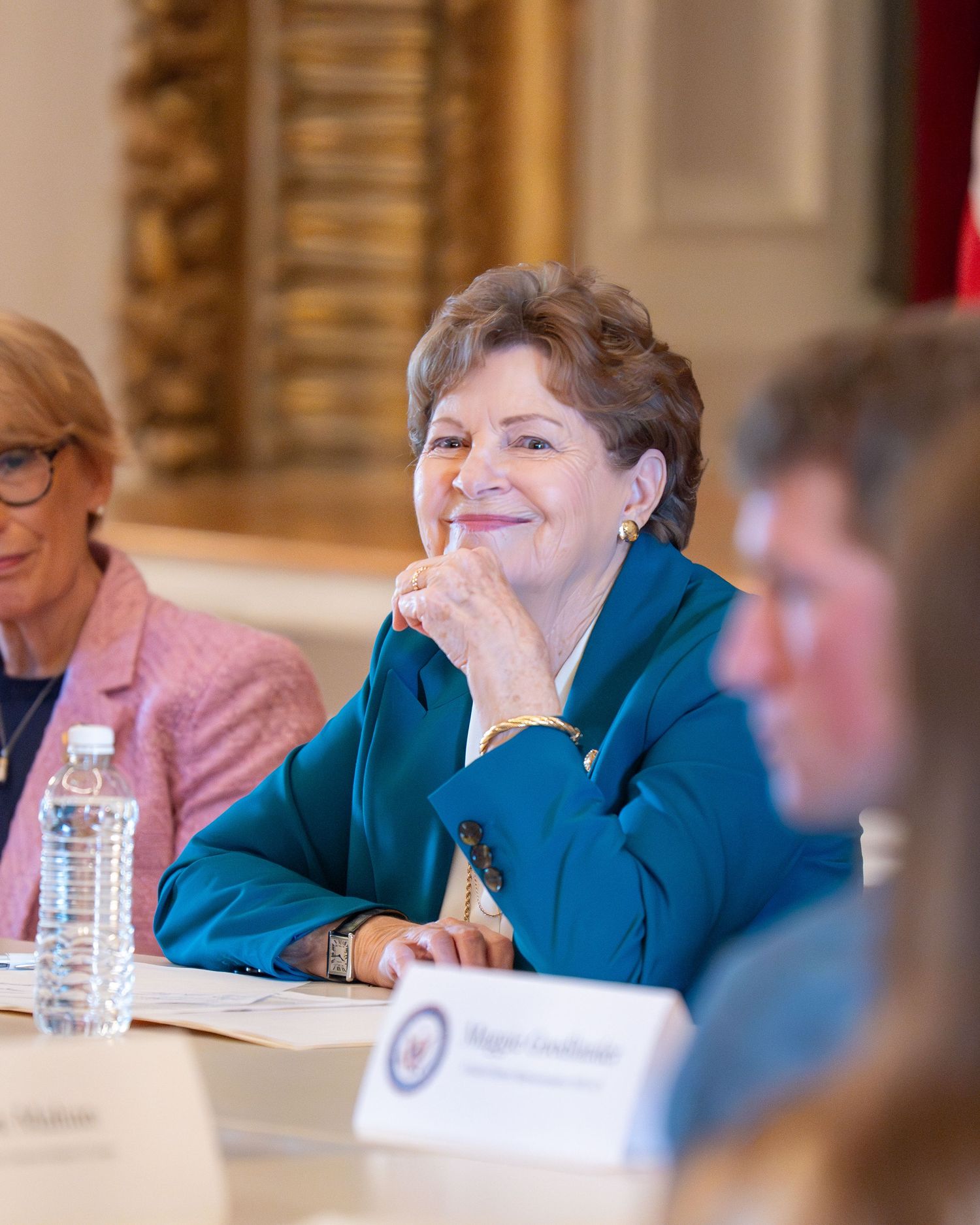 Congresswoman in teal blazer smiles at a formal meeting table with name placards and water bottles at a roundtable