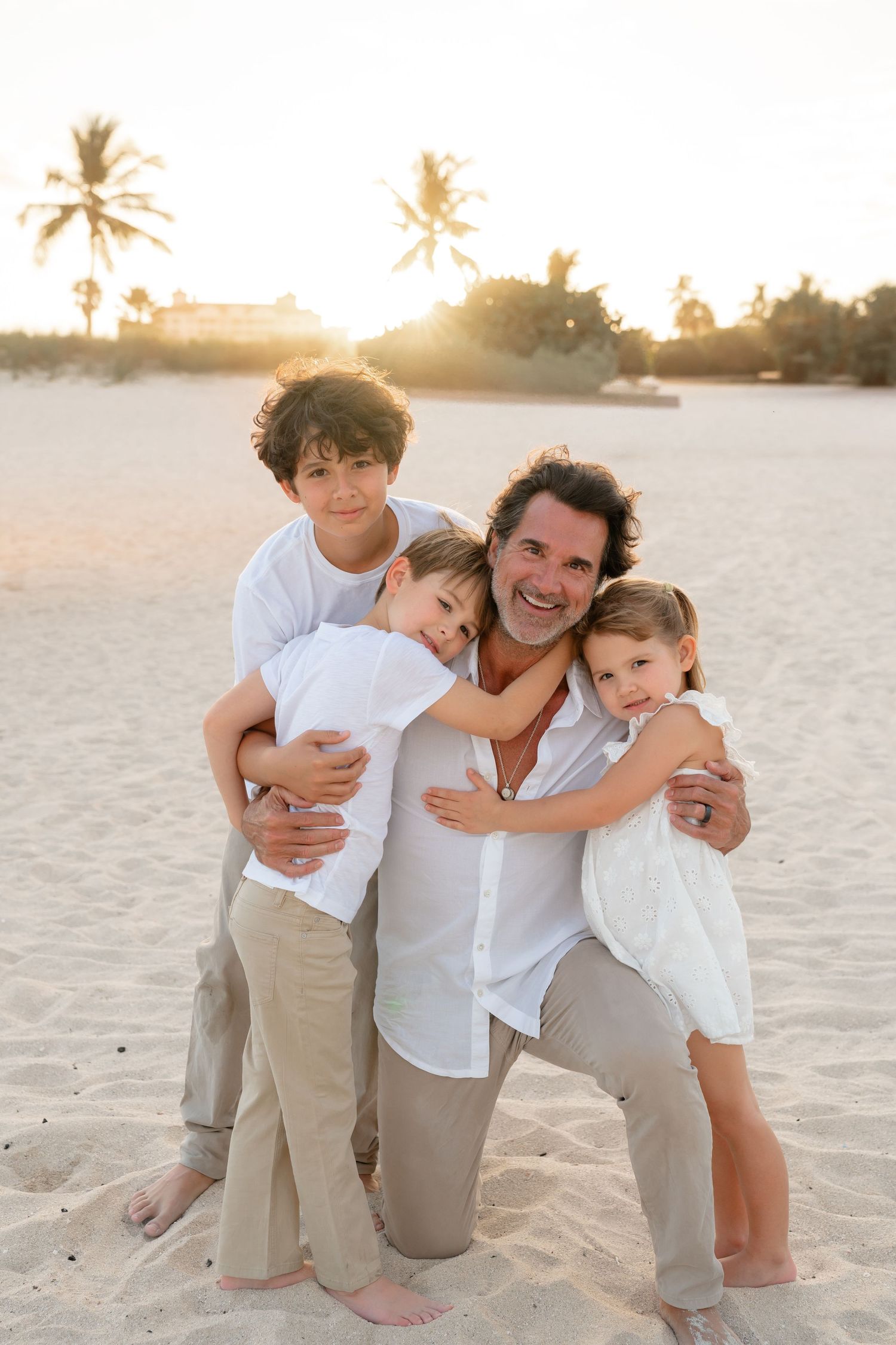 Father kneeling on sandy beach hugging two young sons, smiling outdoors near palm trees at sunset.