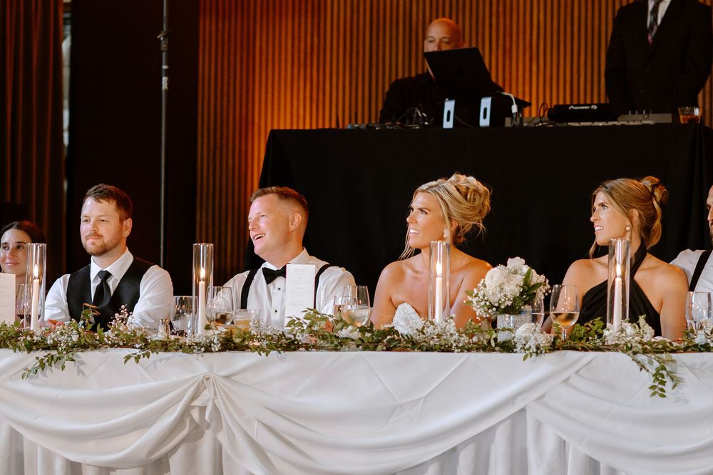 Wedding party seated at a decorated head table with white draping, floral arrangements, and candles during a reception.