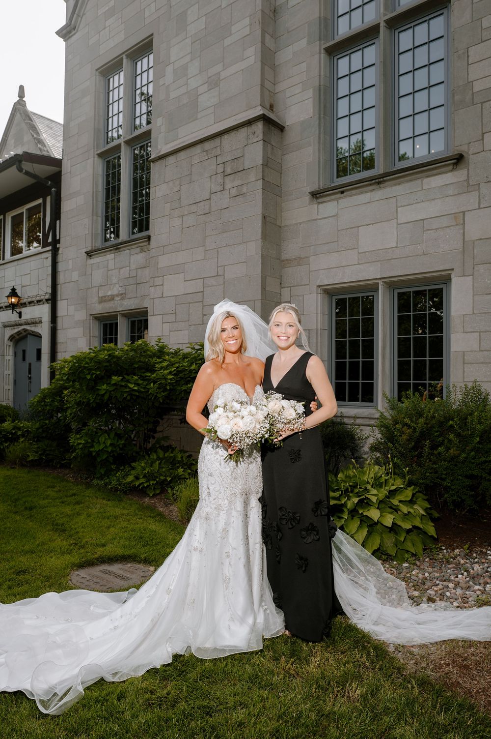 Bride in lace gown with veil poses with bridesmaid in black dress outside stone mansion wedding venue.