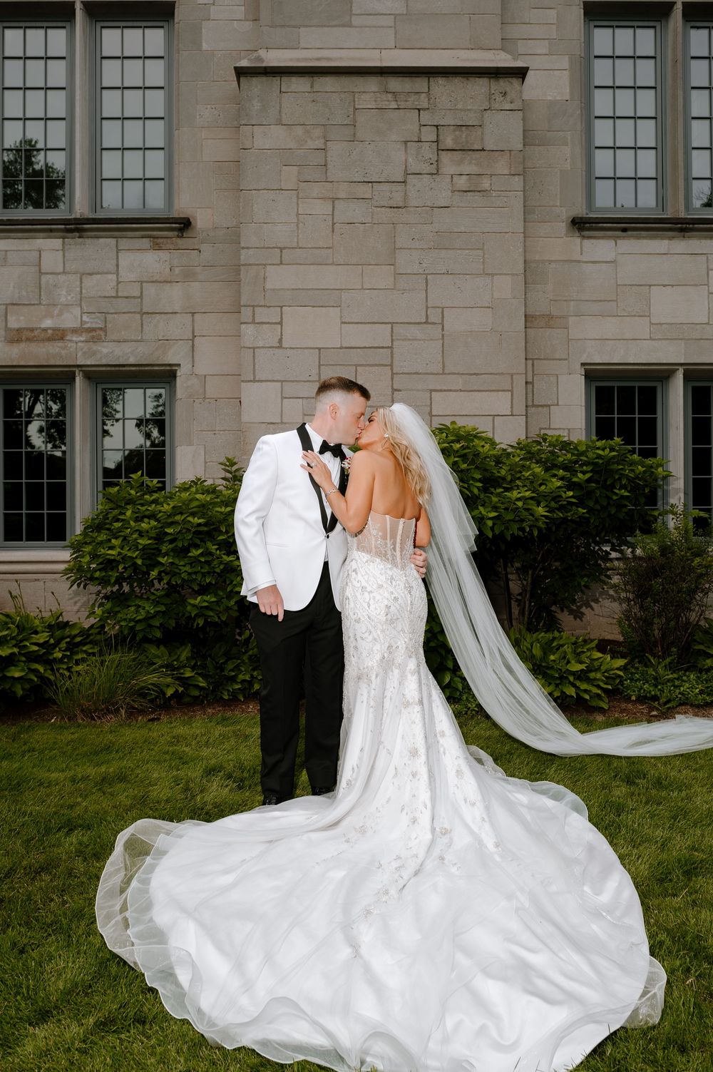 Bride and groom share a romantic kiss outdoors in front of a stone mansion, her lace gown and veil flowing gracefully.