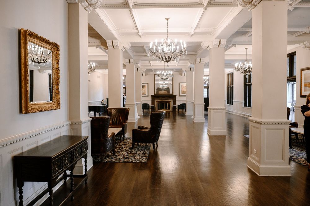 Elegant hotel lobby with white columns, chandeliers, dark hardwood floors, and classic decor including a gold ornate mirror.