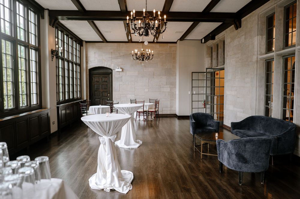 Elegant venue cocktail hour space with high ceilings, dark wood beams, chandelier, cocktail tables, and velvet lounge chairs.