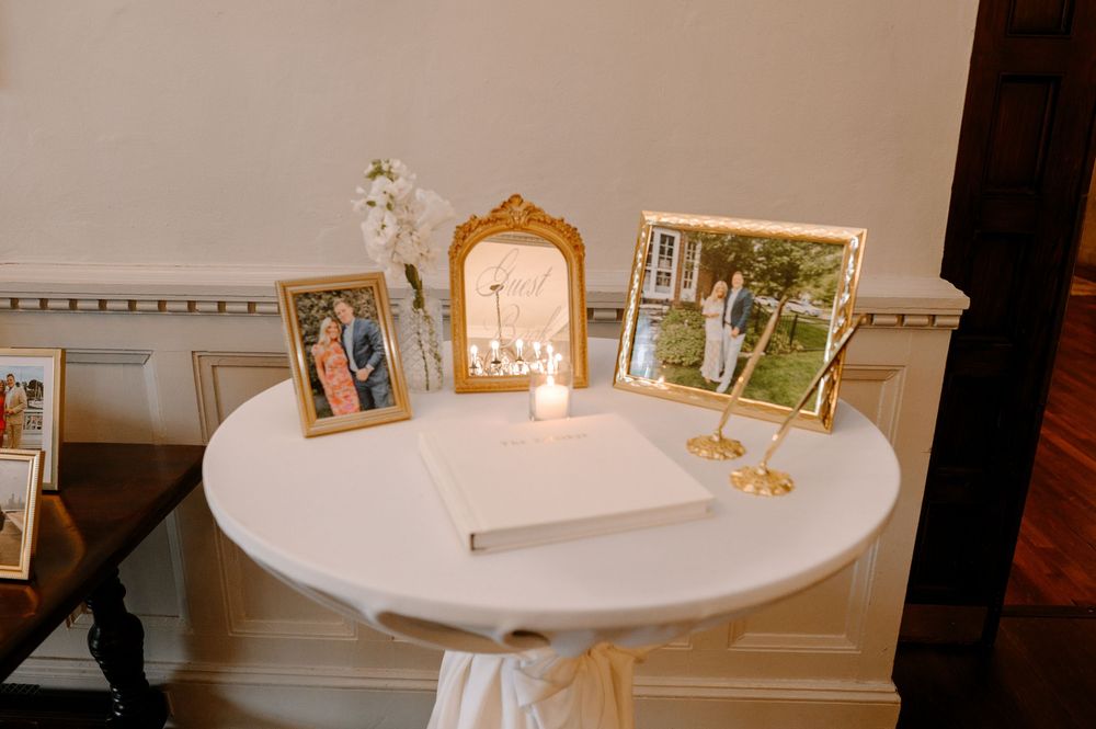 White round table with gold-framed family photos, a guest book, candle, and floral arrangement at a wedding reception.