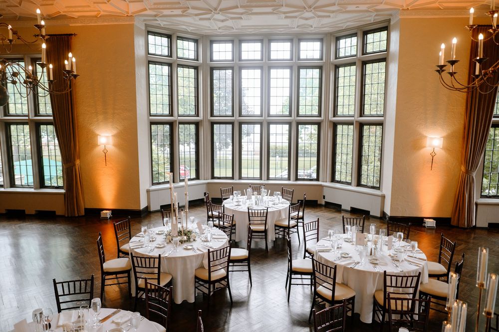 Elegant wedding reception hall with tall leaded glass windows, round tables with white linens, and warm amber uplighting.