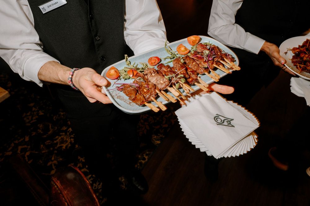 Waiter serving skewered appetizers on a white tray at a catered event, wearing a black vest and white shirt.