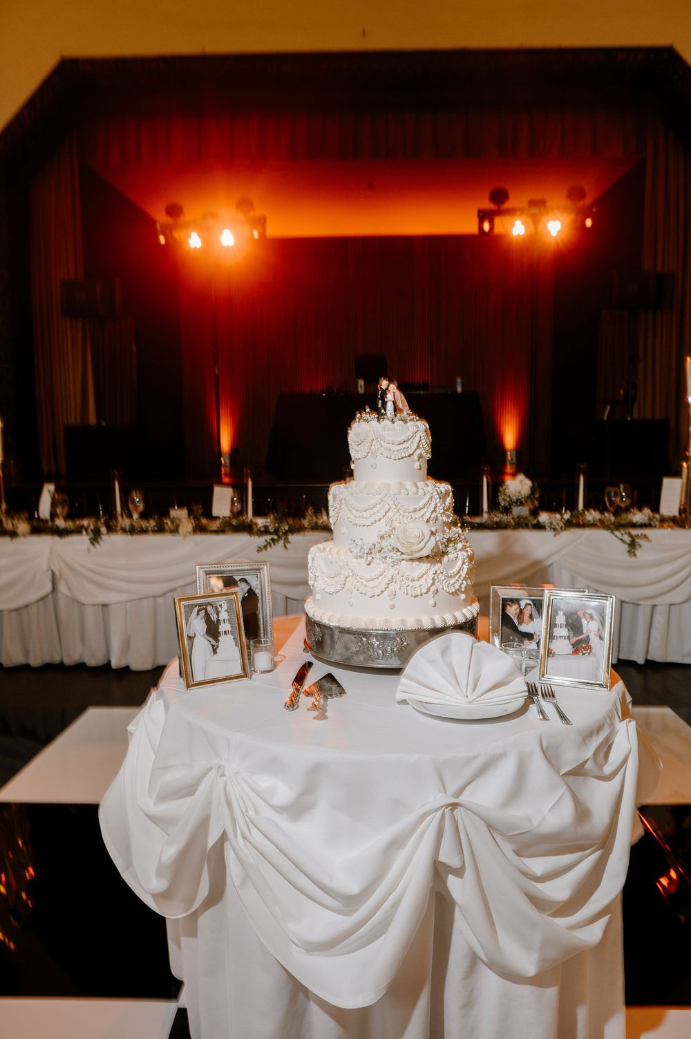 Elegant white tiered wedding cake with floral topper displayed on draped table with framed photos on black and white dance floor.