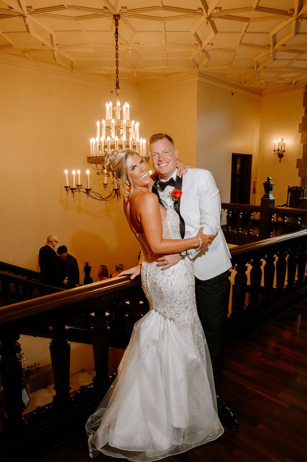 Bride and groom embracing on grand staircase under chandelier, bride in fitted beaded gown, groom in white tuxedo jacket.