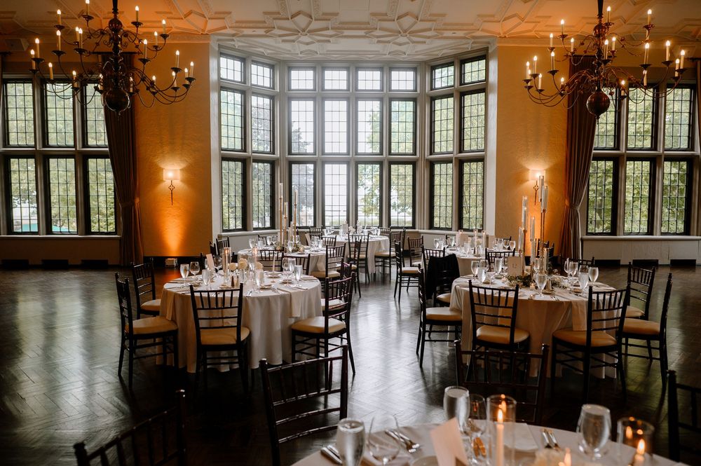 Elegant wedding reception hall with round tables, chiavari chairs, chandeliers, and large arched windows overlooking lush greenery.
