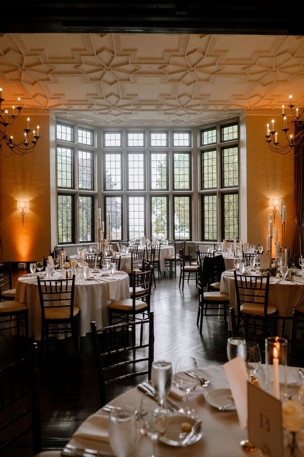 Grand wedding reception room featuring ornate ceiling details, bay windows, candlelit tables, and chiavari chairs.