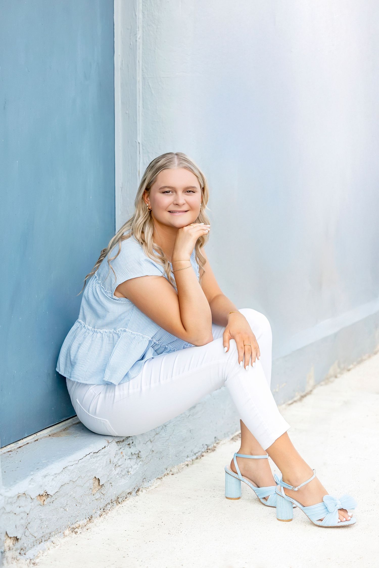 Smiling blonde teen girl in light blue top and white pants sitting against blue wall outdoors.