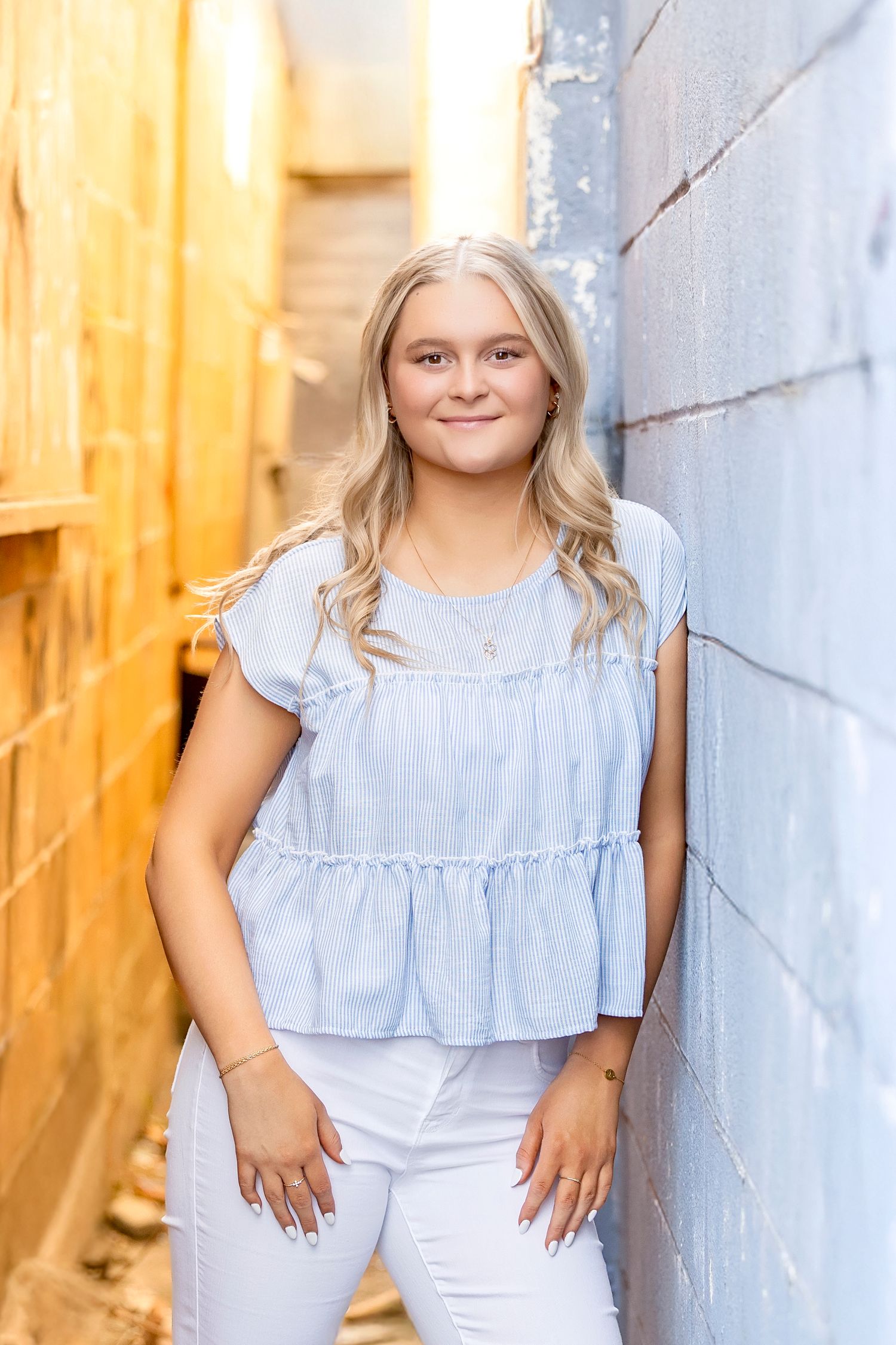 Smiling blonde teen girl in light blue peplum top and white pants leaning against brick wall.