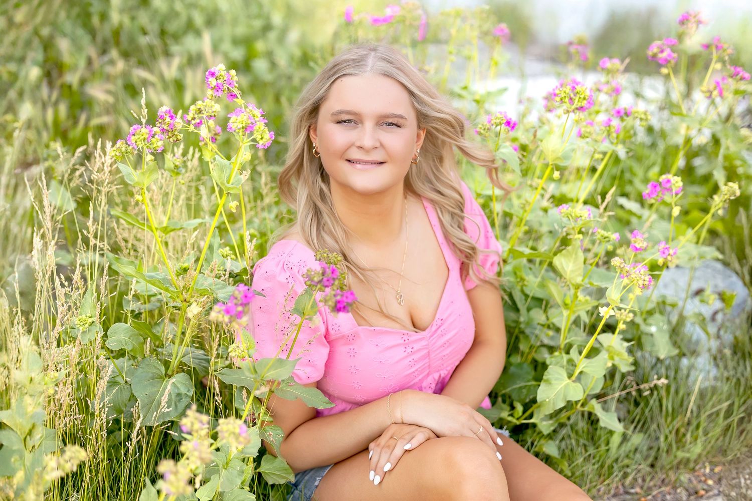 Young blonde woman in pink top sitting among pink wildflowers in a sunny garden.