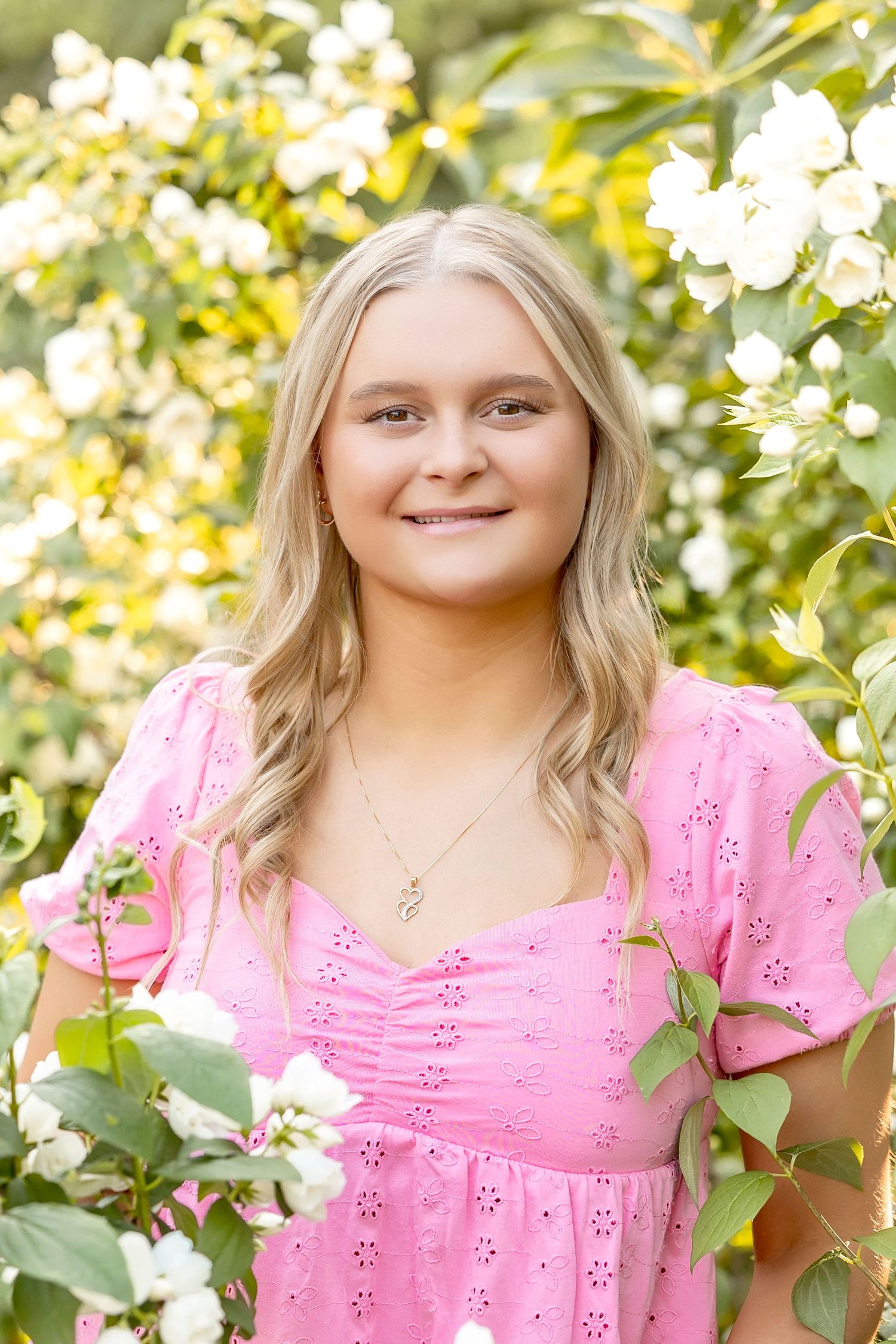 Young blonde woman in pink eyelet dress smiling among white flowering bushes outdoors.