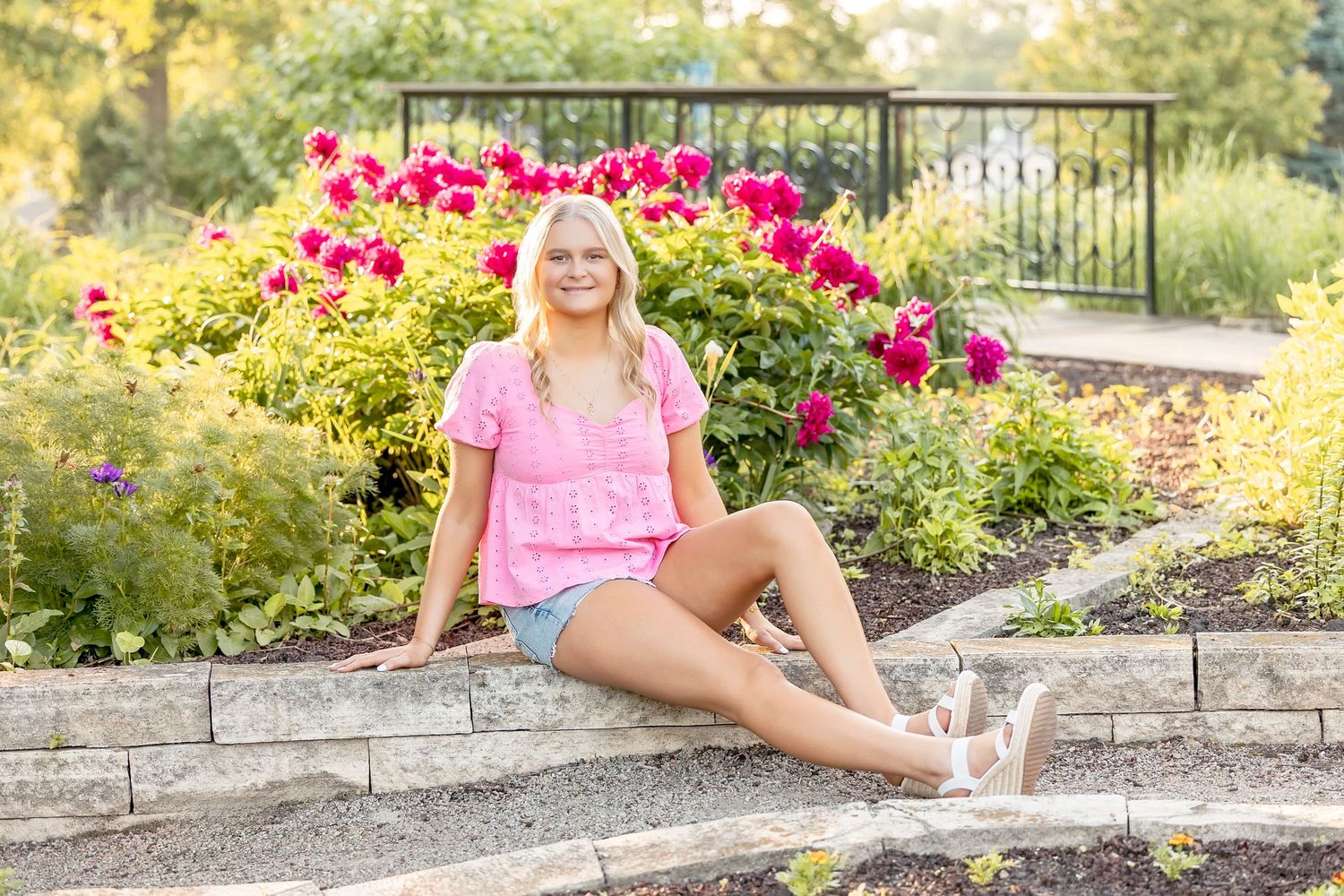 Smiling blonde teen girl in pink top and denim shorts sitting on stone steps surrounded by vibrant red and yellow flowers.
