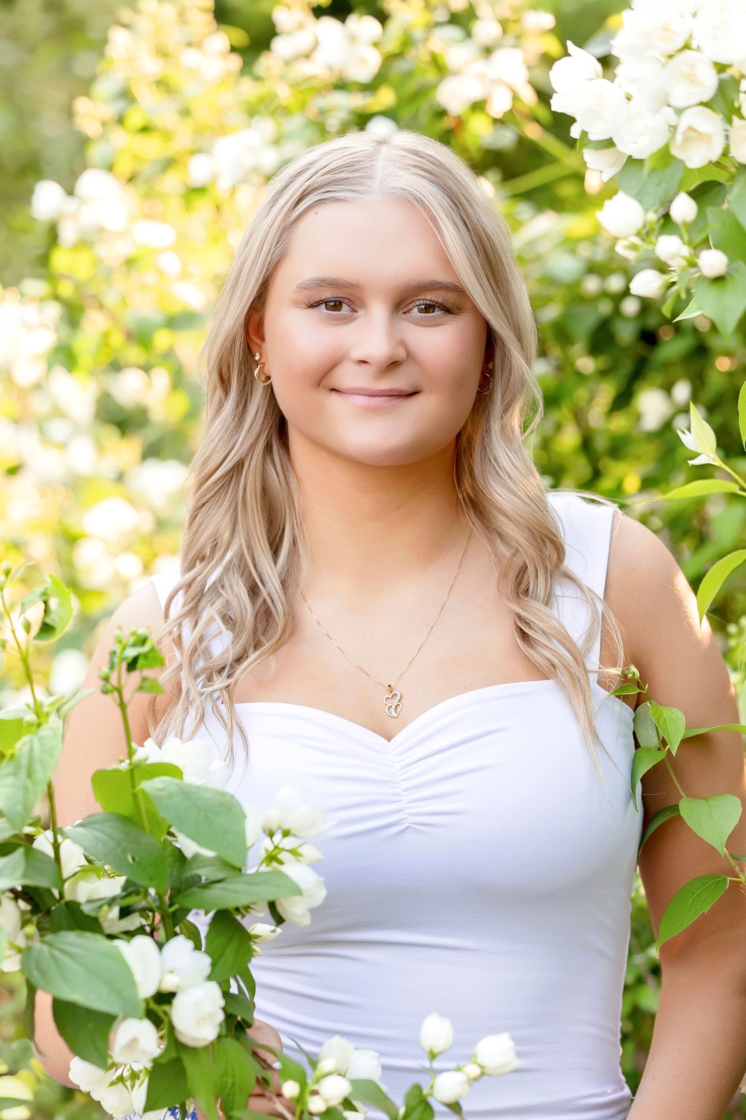 Blonde teenage girl in white top smiling among blooming white flowers in a garden setting.