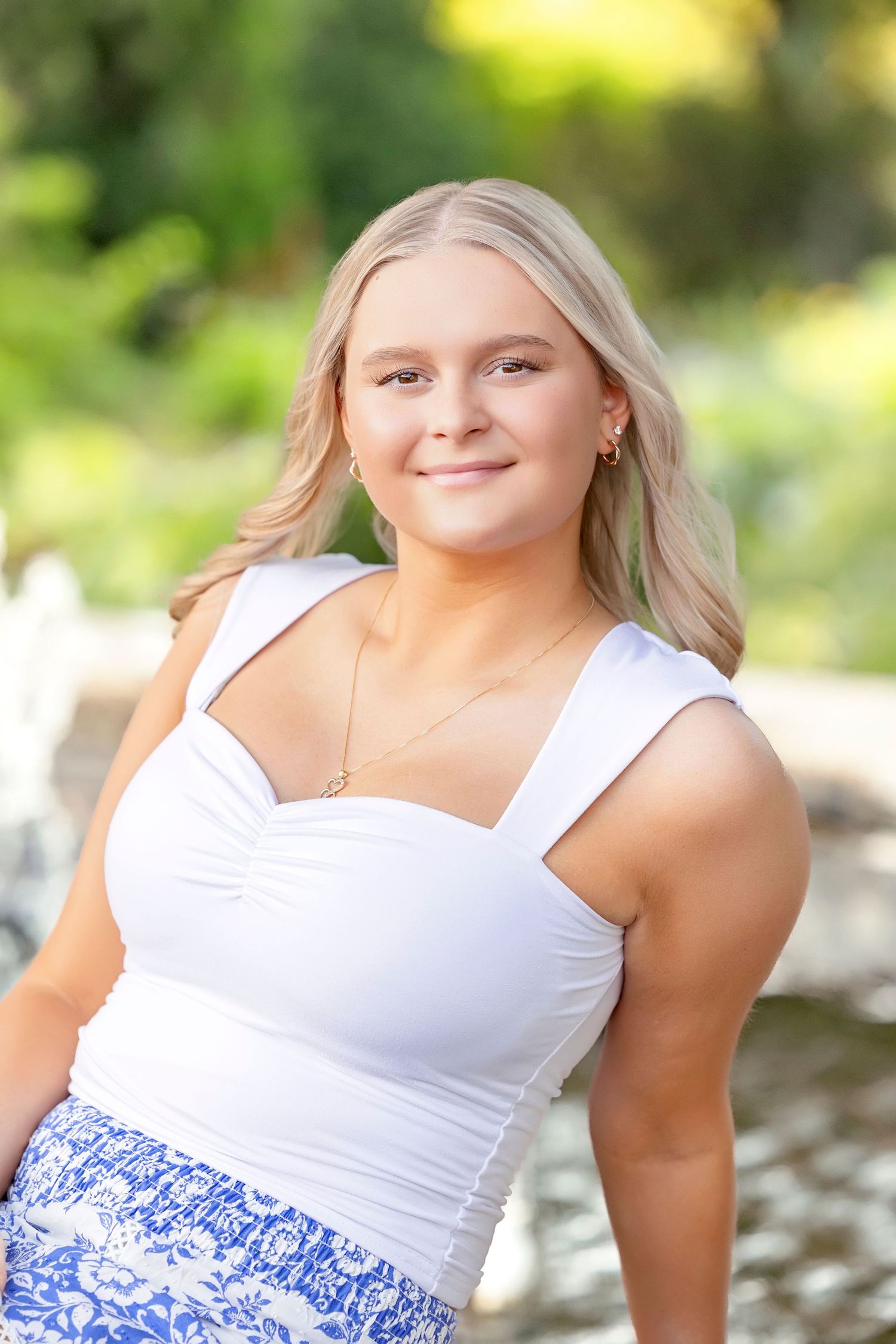 Young blonde woman smiling outdoors in white top and blue floral skirt, posing in a lush green garden setting.