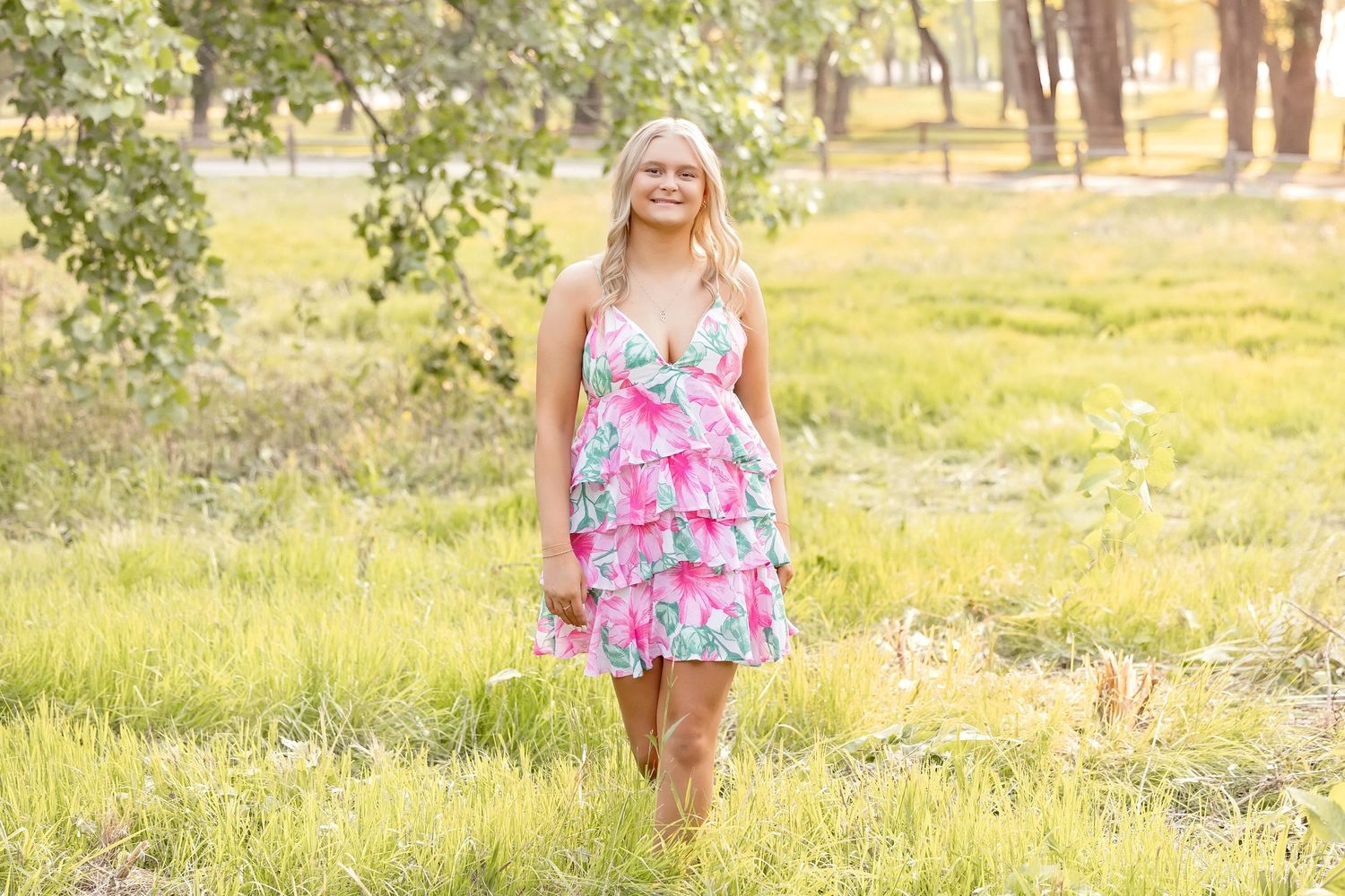 Young blonde girl in colorful pink ruffled dress smiling while walking through a sunny green field.