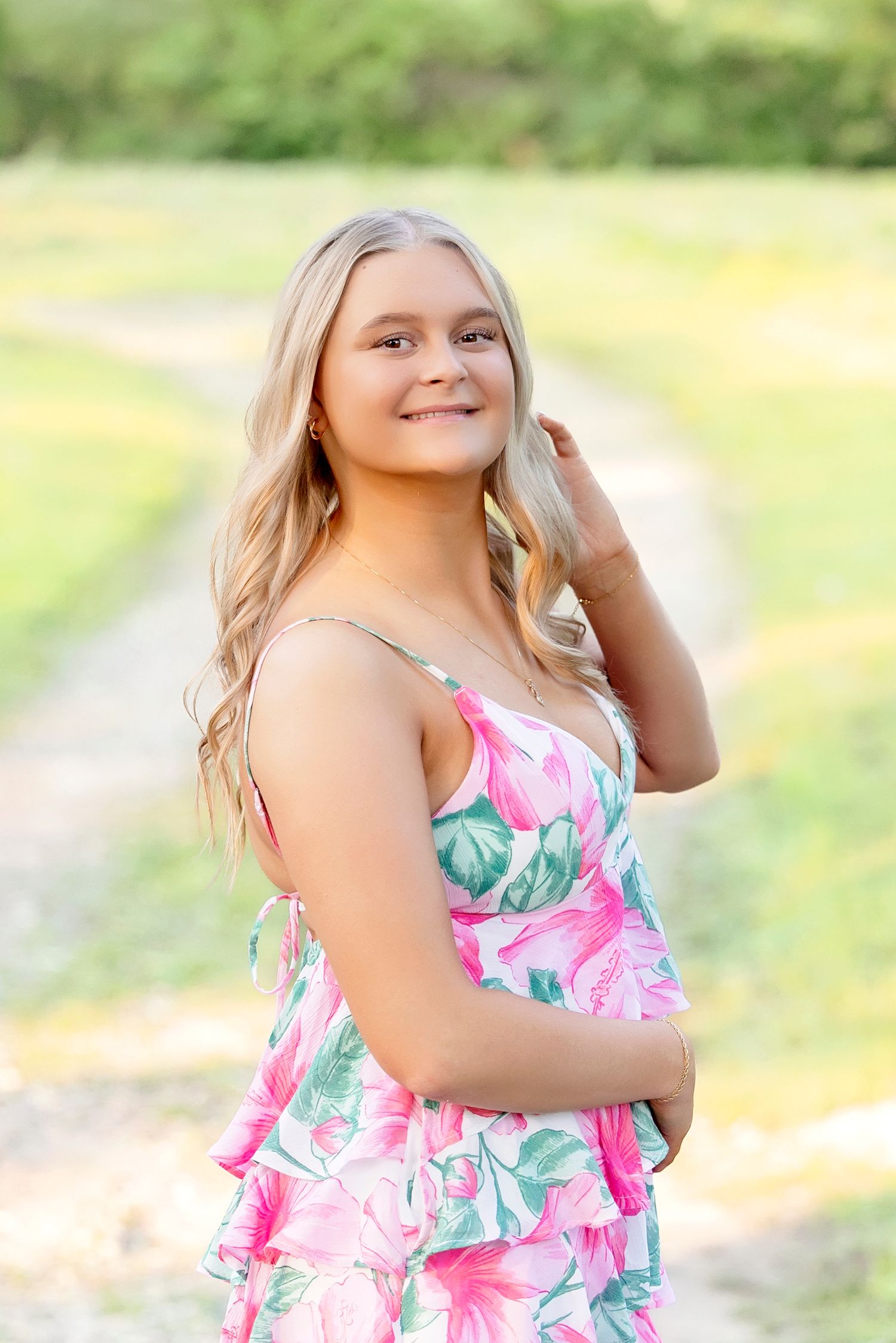 Smiling blonde teen girl in pink floral dress posing outdoors on a sunny day with green trees in background.