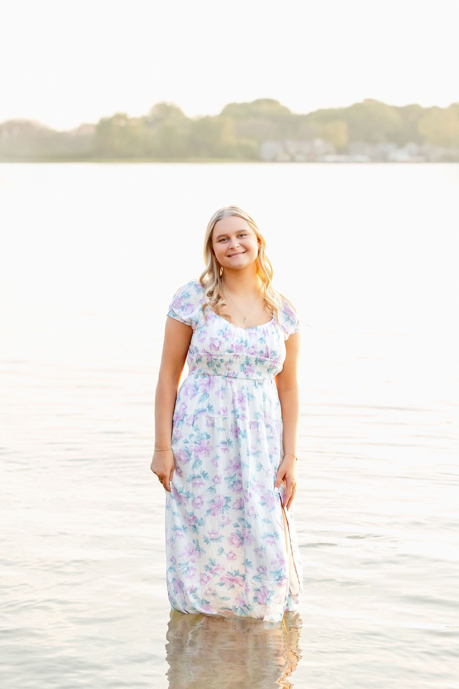 Smiling blonde teen girl in floral maxi dress standing in shallow lake water at golden hour.