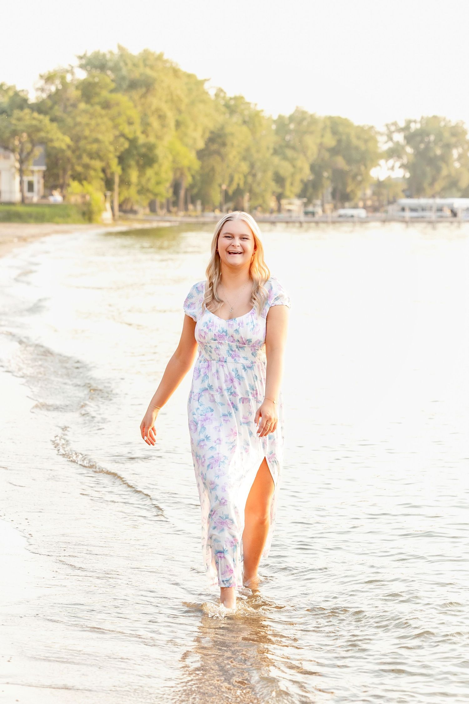 Laughing blonde woman in floral maxi dress walking barefoot along a sunny lakeside beach.
