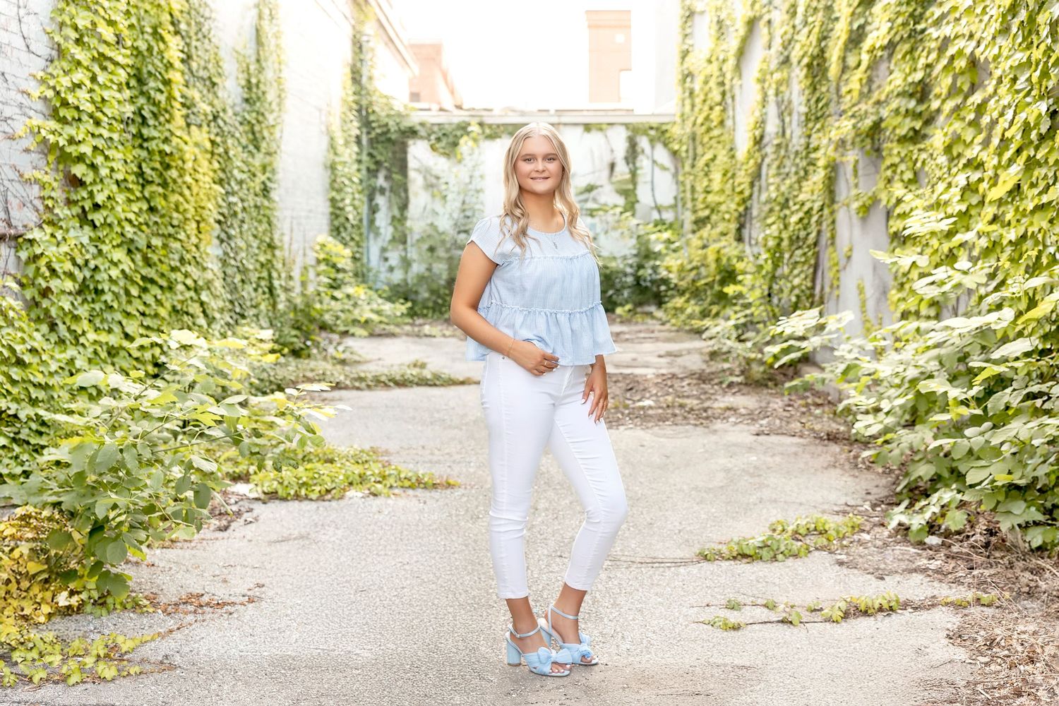 Woman in white jeans and blue top poses on a garden path surrounded by lush green ivy-covered walls.
