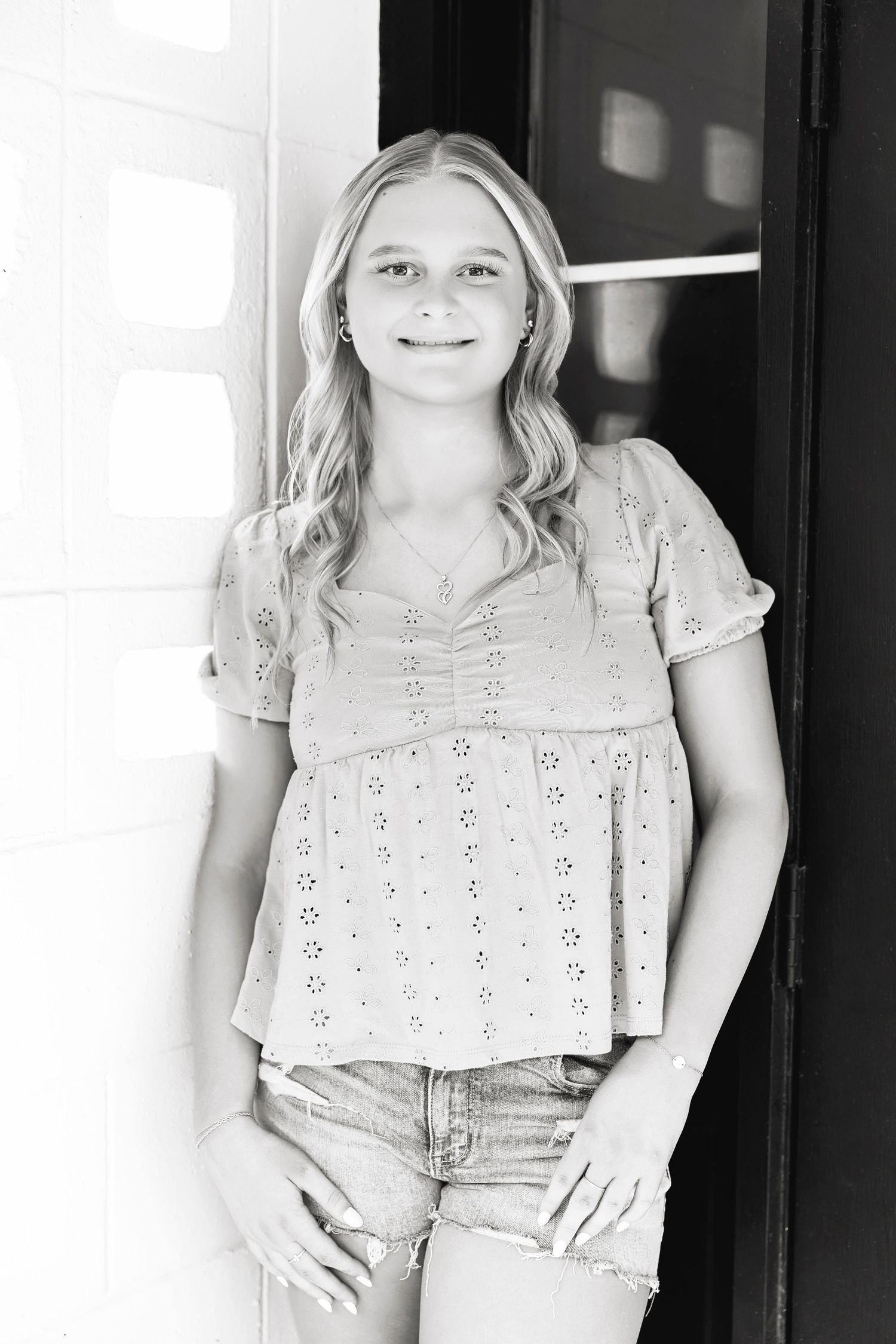 Black and white portrait of a blonde teenage girl in eyelet top and denim shorts by a doorway.