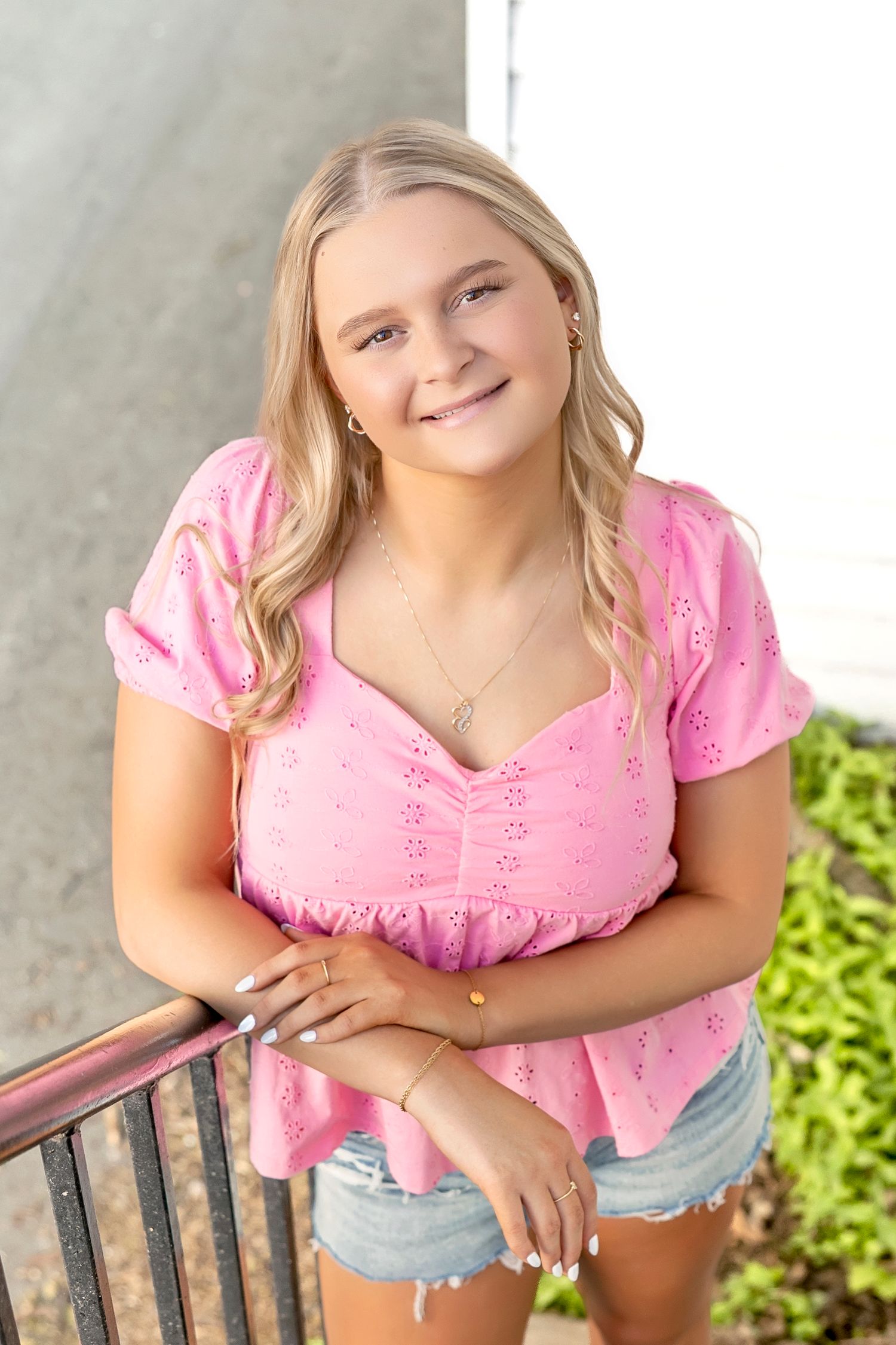 Smiling teenage girl in pink eyelet top and denim shorts leaning on a railing outdoors.