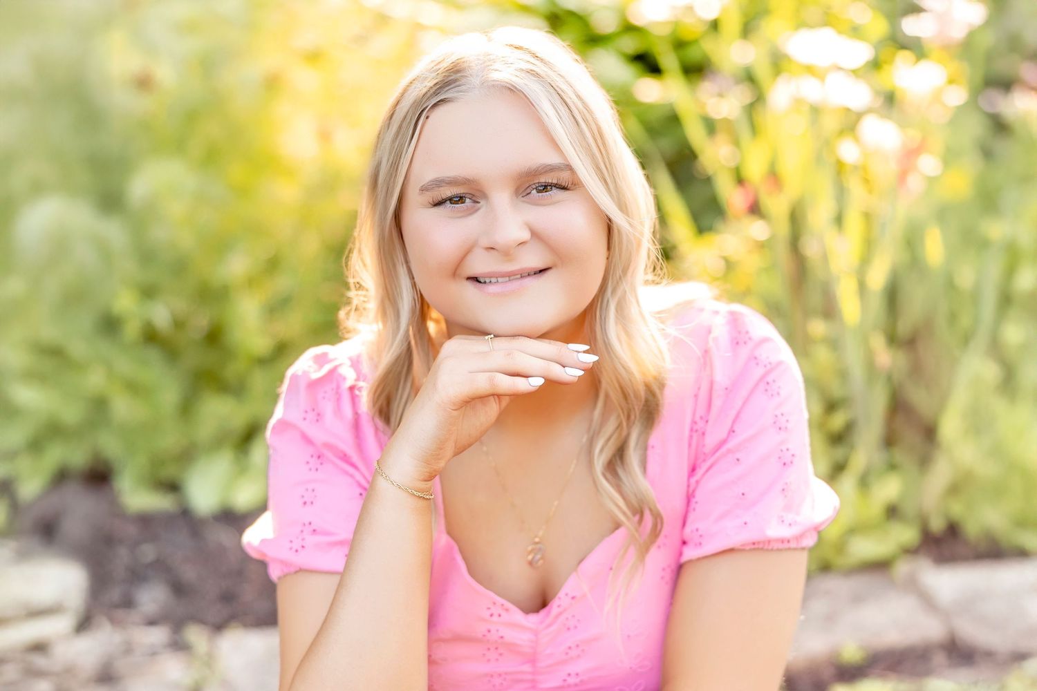 Smiling young blonde woman in pink top posing outdoors with lush green garden background in natural sunlight.