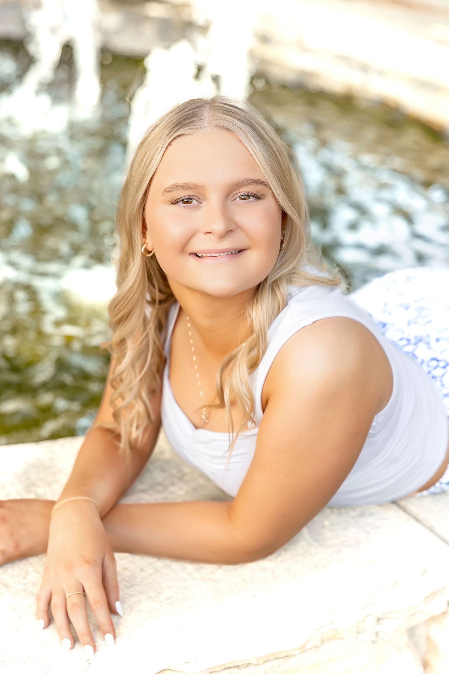 Smiling blonde teenage girl in white top posing by a fountain outdoors in warm sunlight.