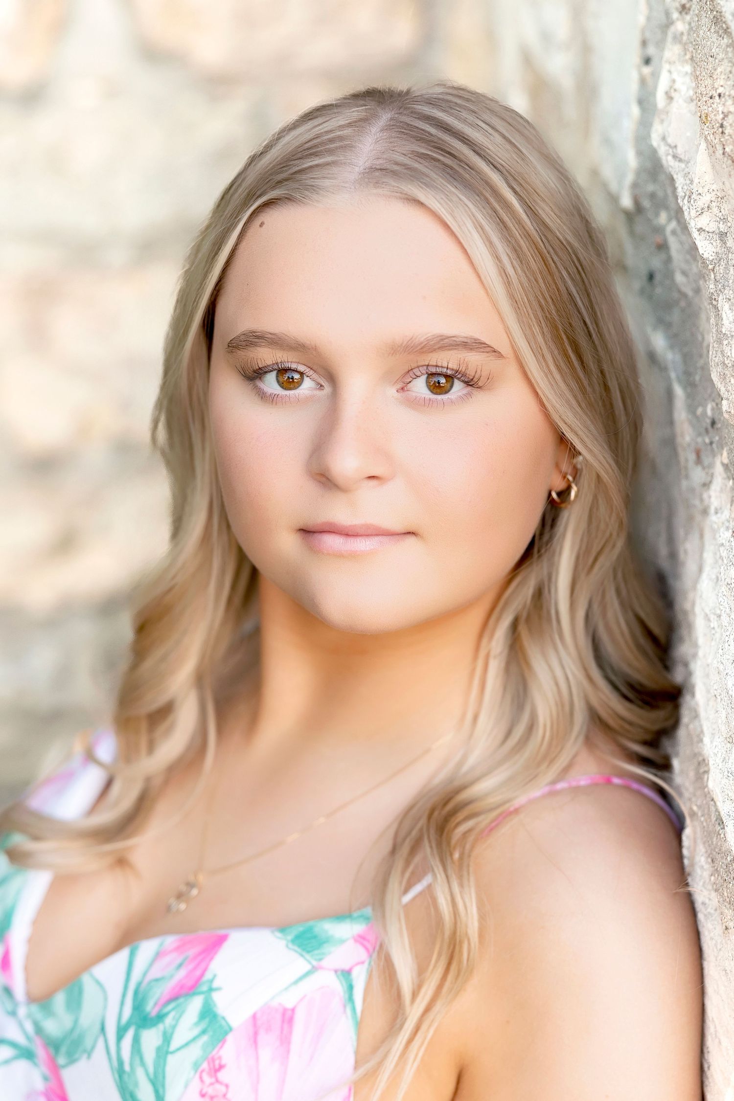 Blonde teenage girl with brown eyes wearing a floral dress, posing against a stone wall outdoors.