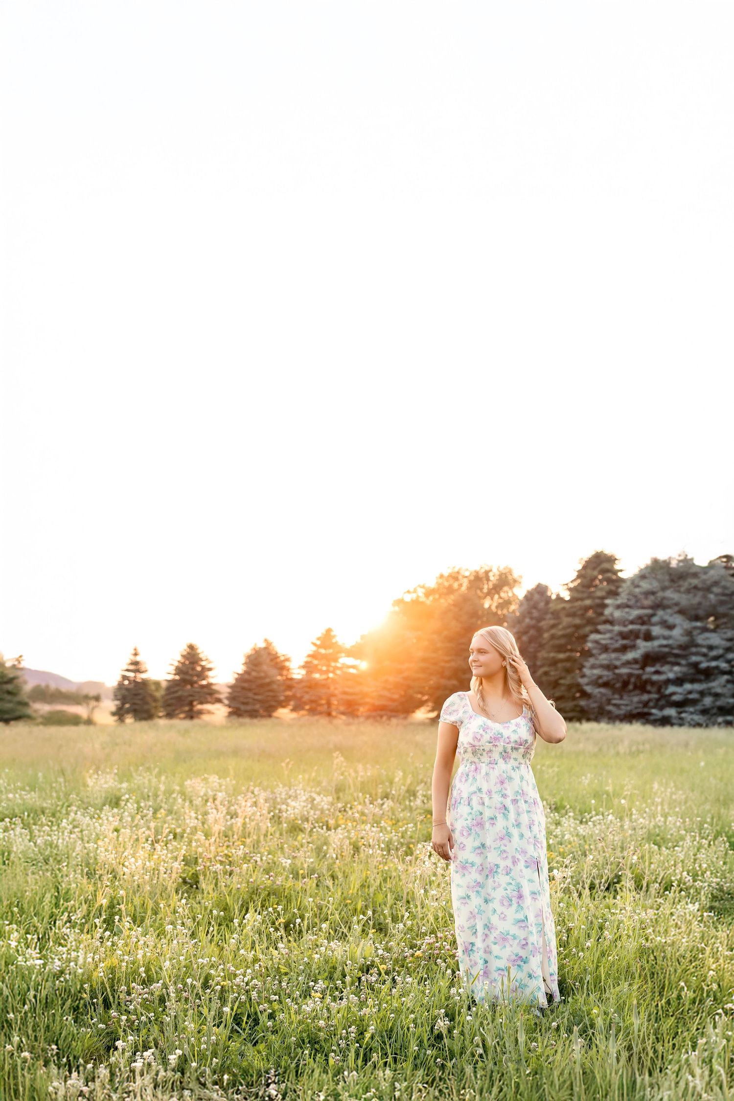 Woman in floral maxi dress standing in sunlit meadow with wildflowers and trees at golden hour sunset.