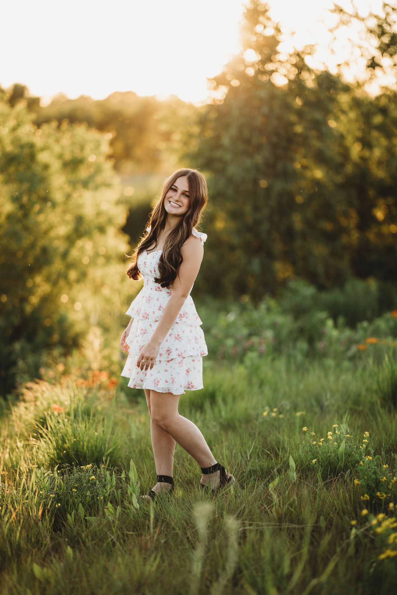 Young woman in white floral dress smiling in sunlit green meadow during golden hour.