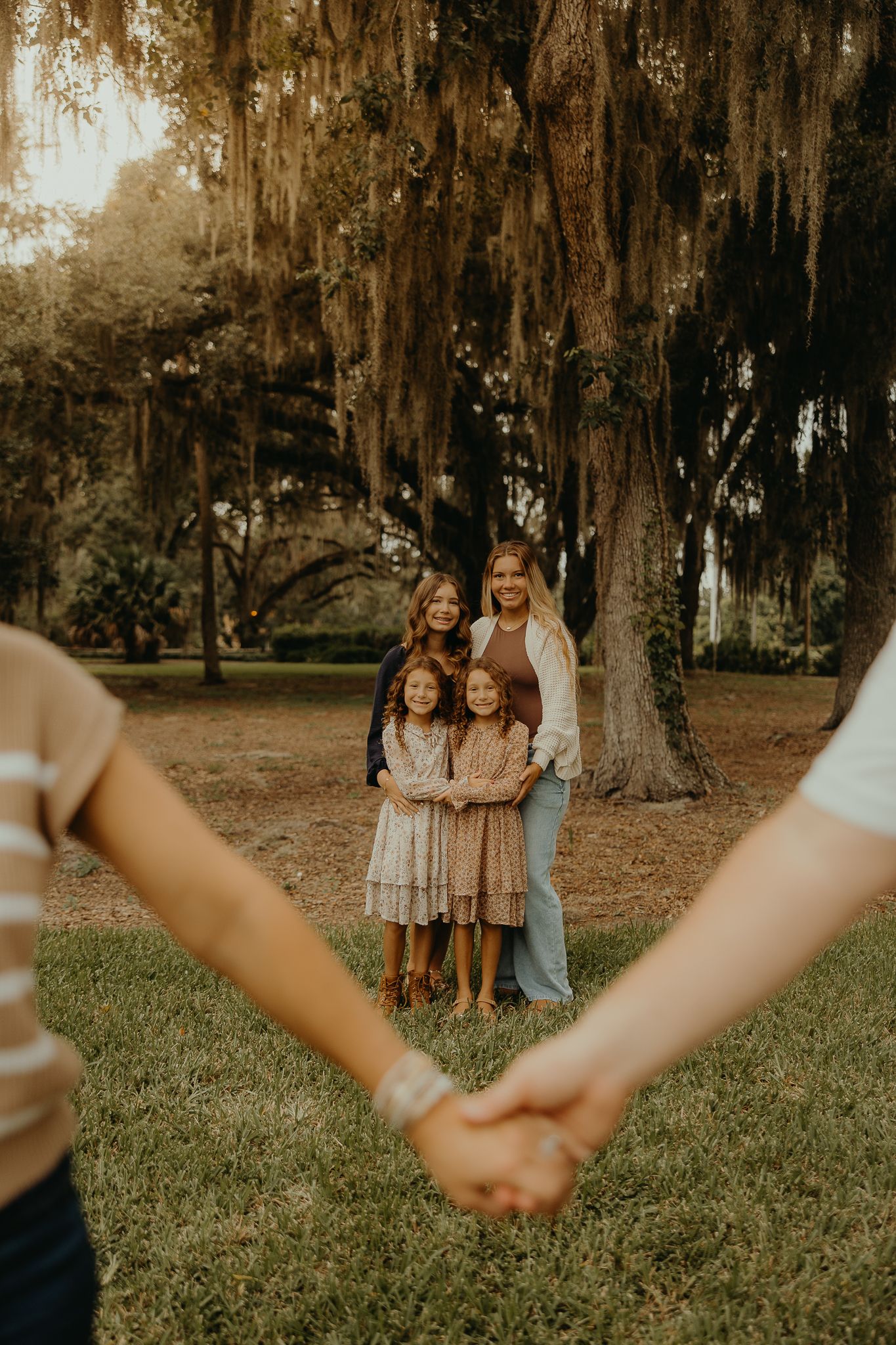 Two adults and two children hold hands in a lush park under moss-draped oak trees during golden hour.