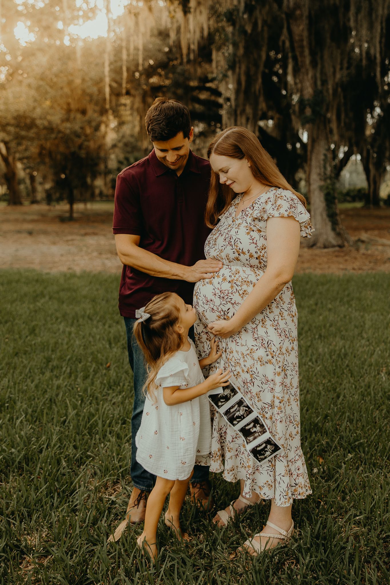 Two adults and two children hold hands in a lush park under moss-draped oak trees during golden hour.