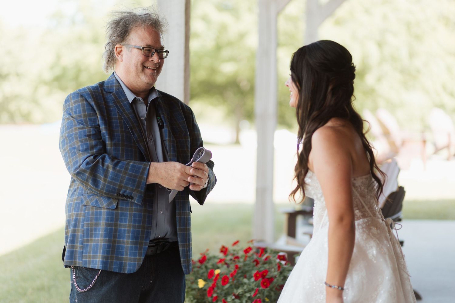 A man in a blue plaid jacket converses with a bride in a white dress outdoors near red flowers.