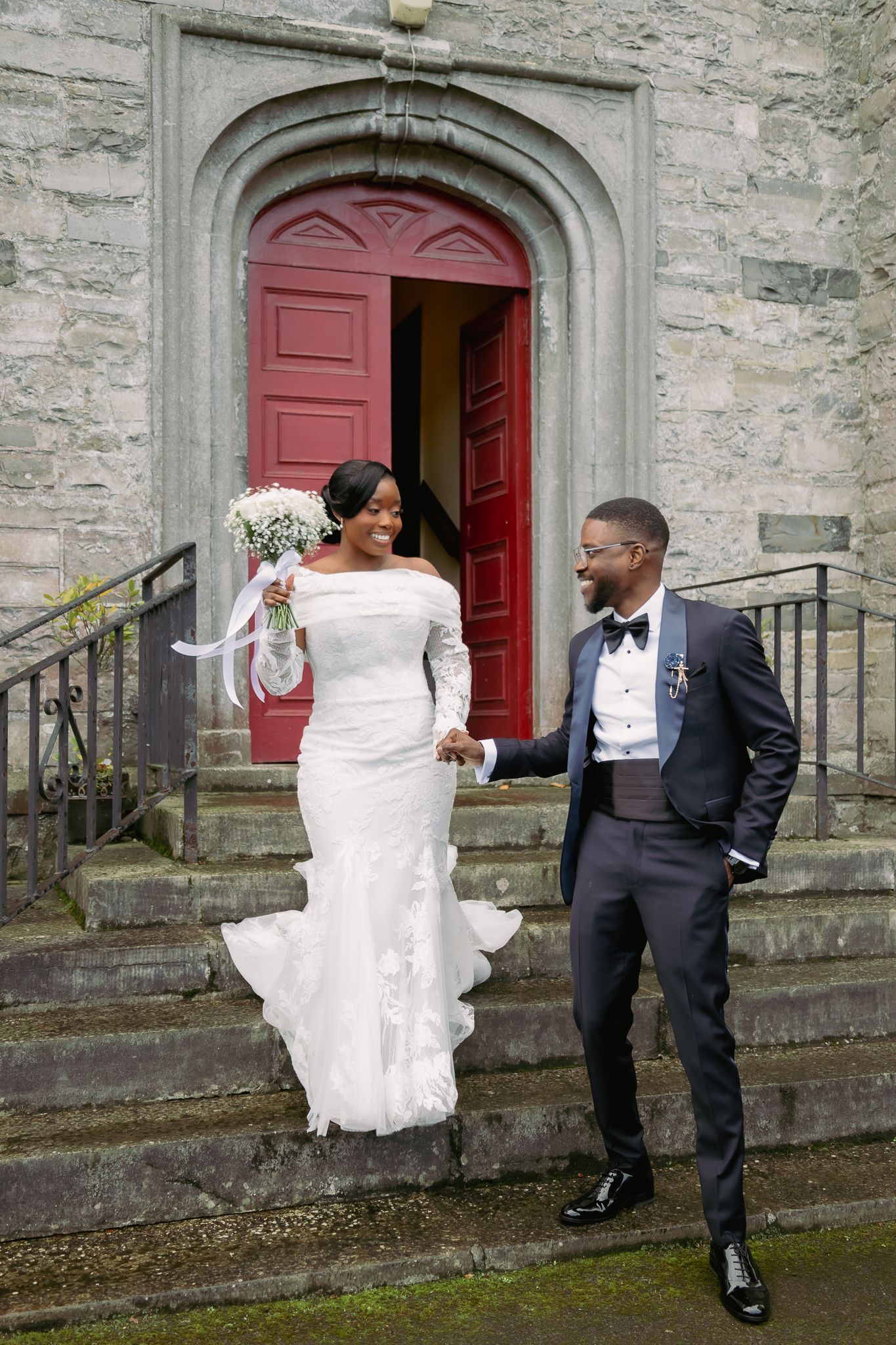 Bride and groom walking down church steps, bride in white gown holding bouquet, groom in navy suit, smiling.