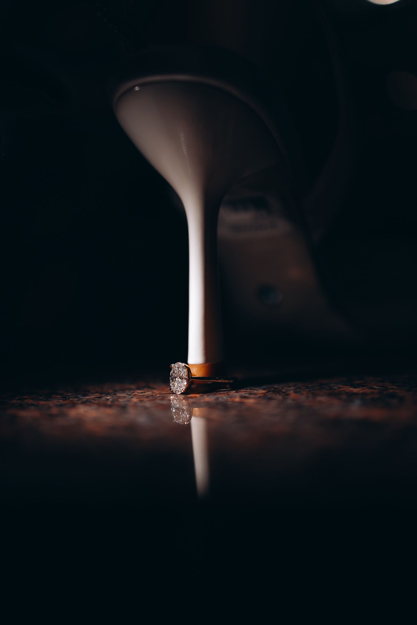 A white stiletto heel resting on a diamond engagement ring on a dark reflective surface.